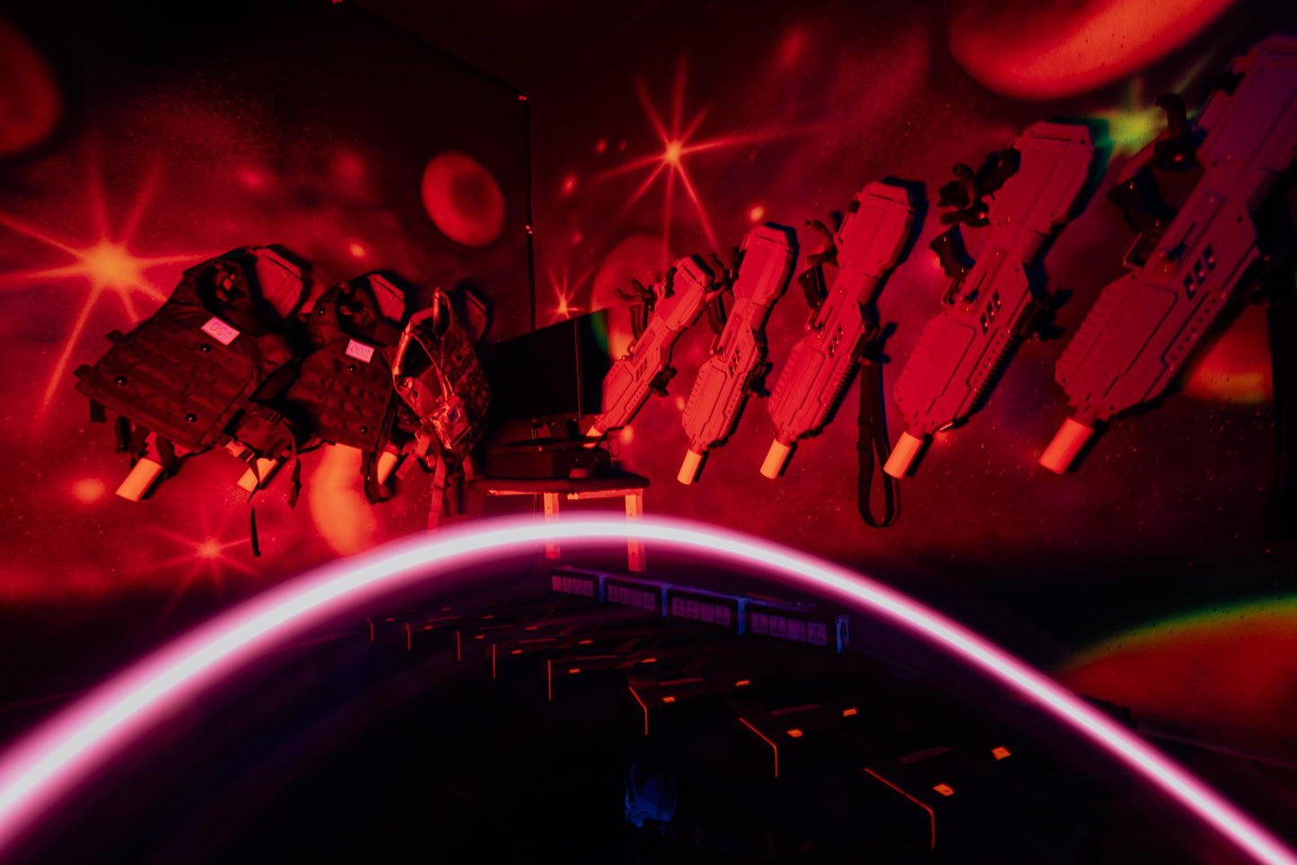Laser tag vests and phasers hang on a wall in a dimly lit, red-lit arena.