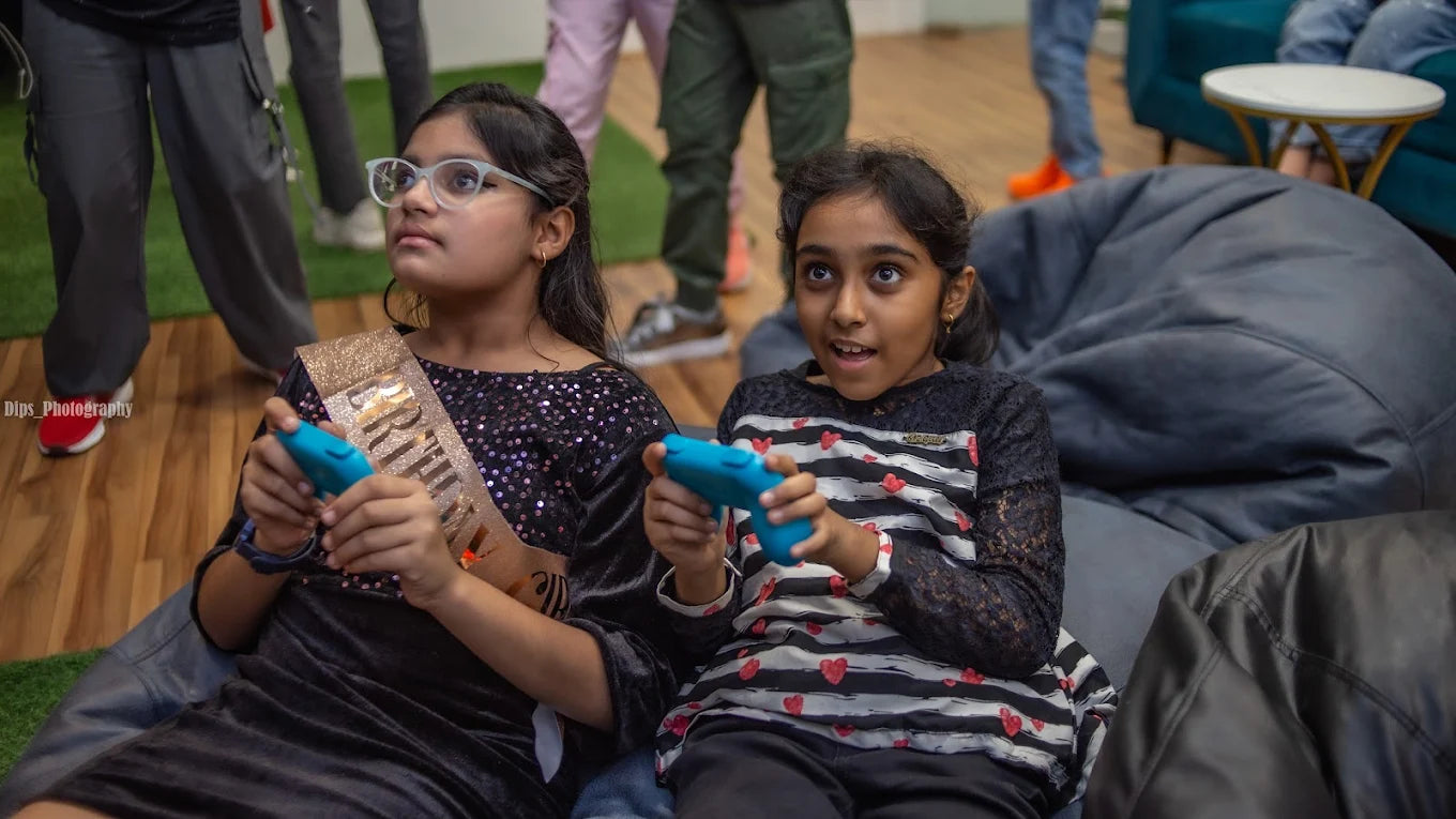 Two young girls play video games at a party, one wearing a birthday sash.