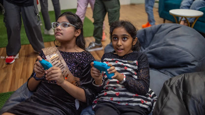 Two young girls play video games at a party, one wearing a birthday sash.