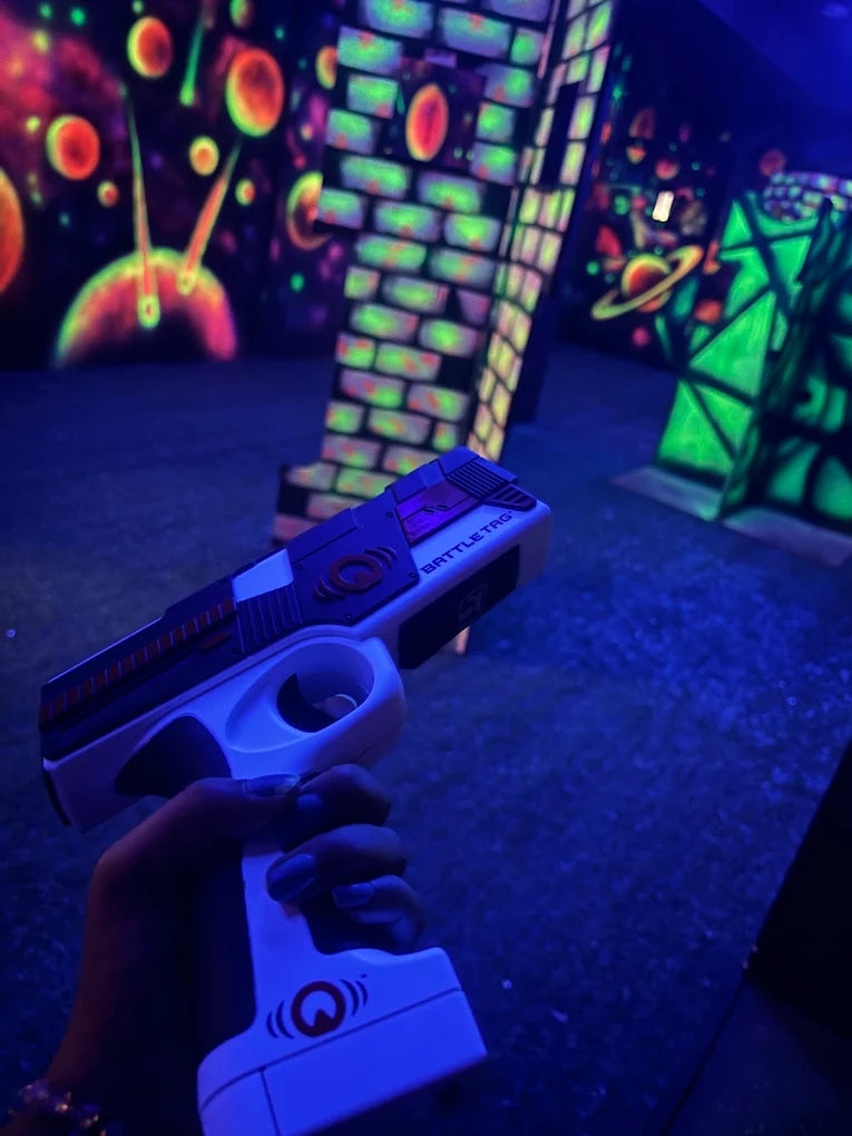 A person holds a laser tag gun in a neon-lit arena with glowing planets and brick walls.
