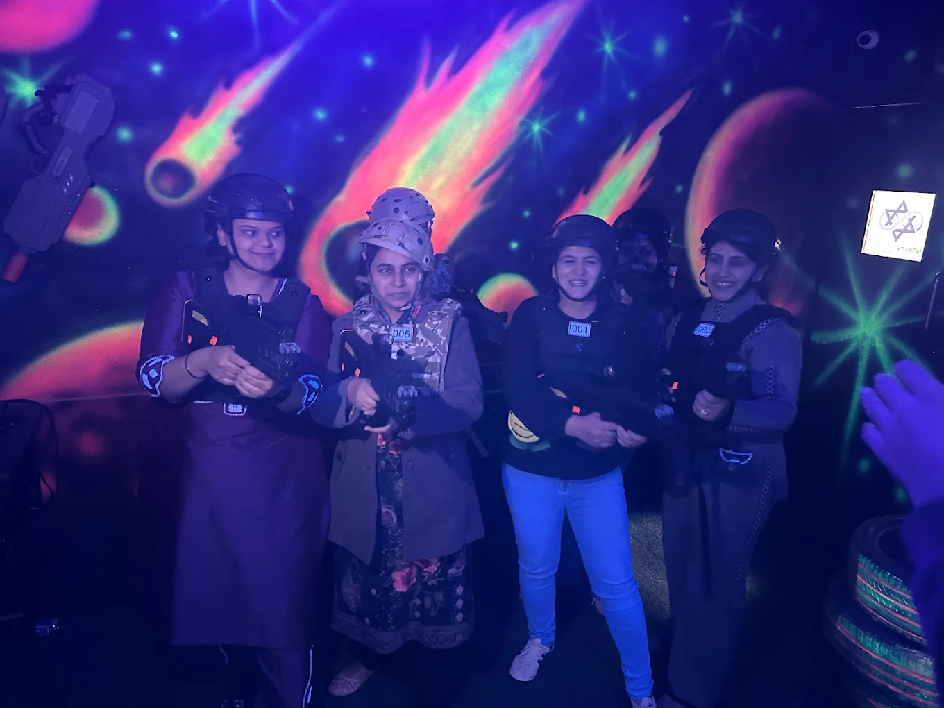 Young women in laser tag gear and helmets pose with their phasers in a neon-lit arena.