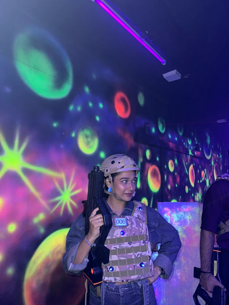 A young woman in a laser tag vest and helmet holds a phaser in a neon-lit arena.
