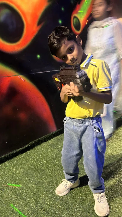 A young boy in a yellow shirt and jeans holds a laser tag gun in a dimly lit arena with colorful murals.