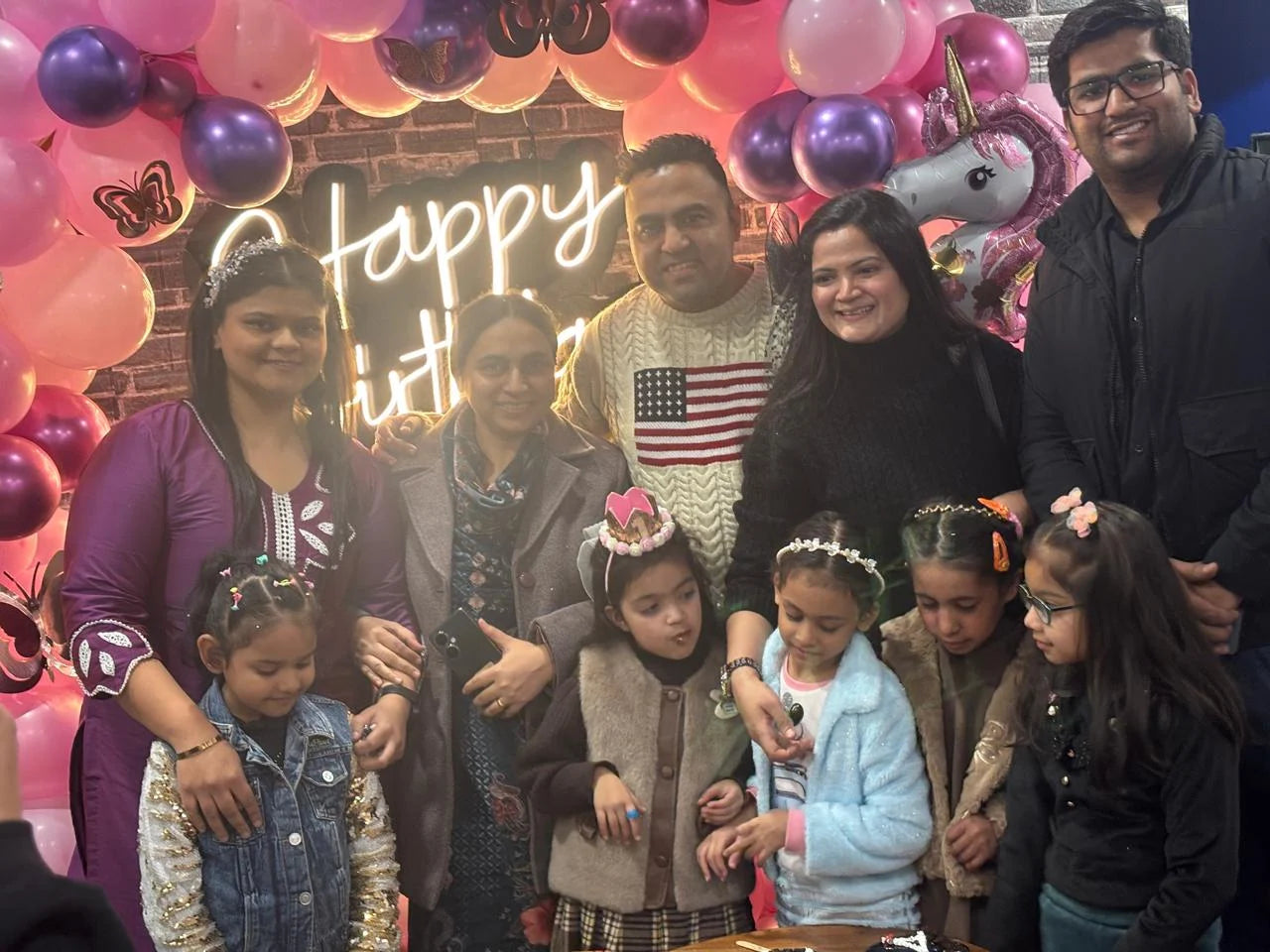A family celebrates a birthday with a cake and balloons, with adults and young children posing for a photo.