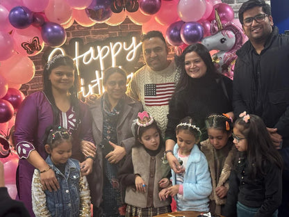 A family celebrates a birthday with a cake and balloons, with adults and young children posing for a photo.
