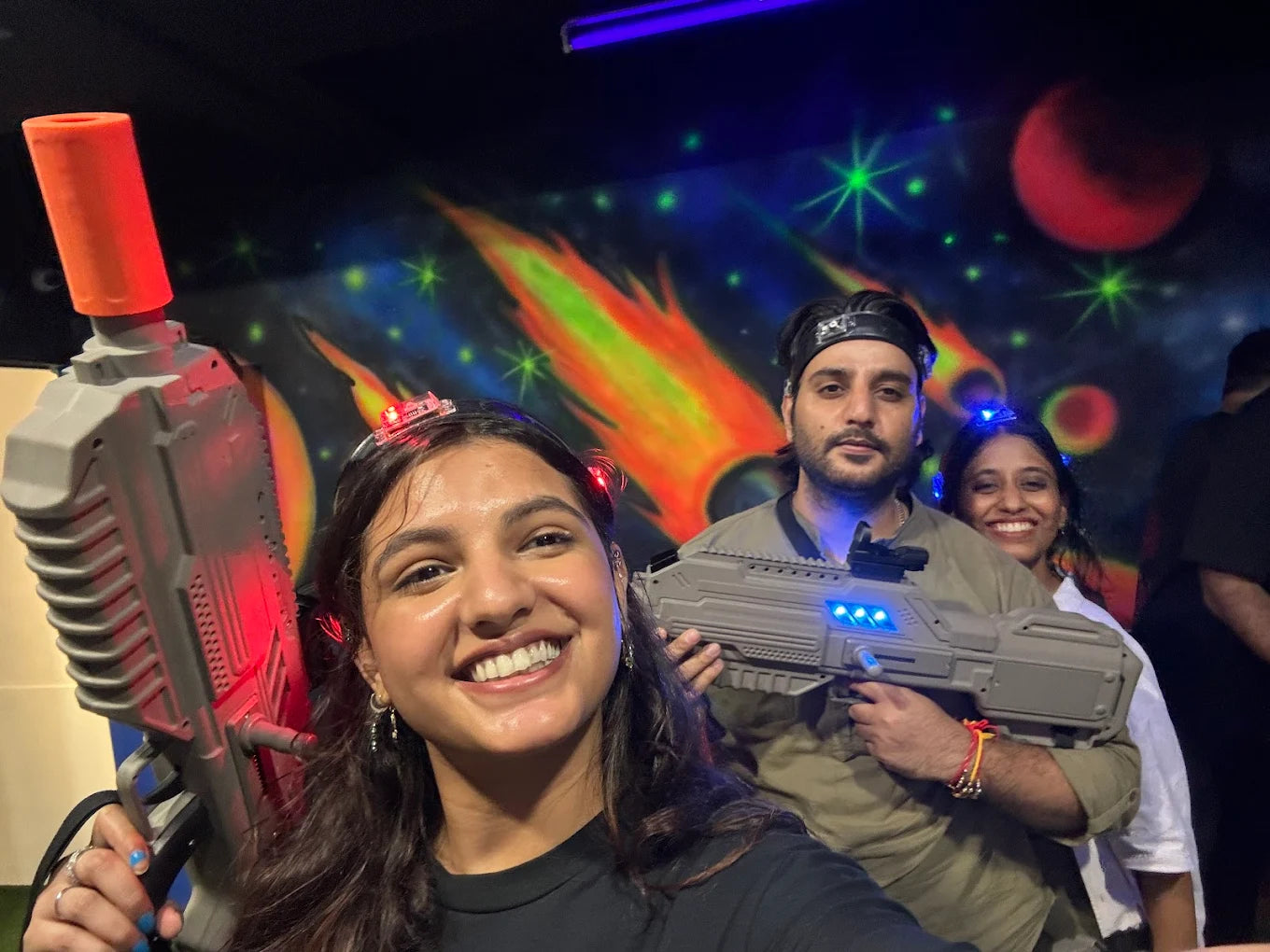 Three young adults play laser tag in a neon-lit arena with a space-themed mural.