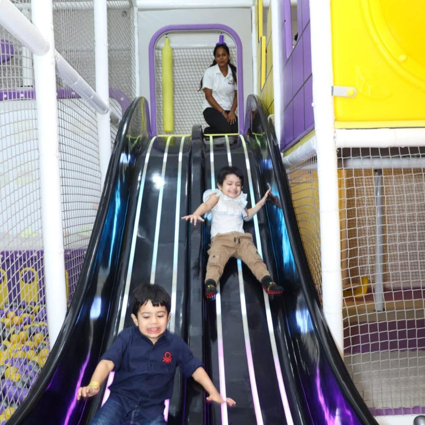 Children sliding down a purple slide in an indoor playground with a woman observing.