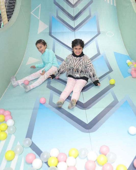 Two girls sliding down a painted indoor slope surrounded by pastel-colored plastic balls at Kidzzilla Fuzion Rajouri Garden
