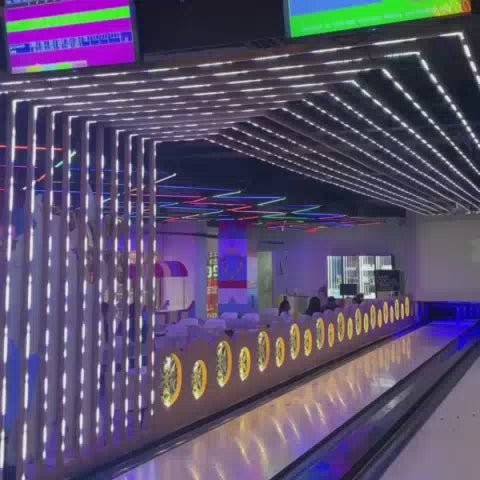 A vibrant arcade buzzes with activity, featuring a bowling alley with neon lights, racing arcade games, and a hammer-smash game. Young adults and children alike enjoy the energetic atmosphere, playing various games like air hockey and claw machines. The modern, brightly lit environment creates a fun and exciting experience for all ages.