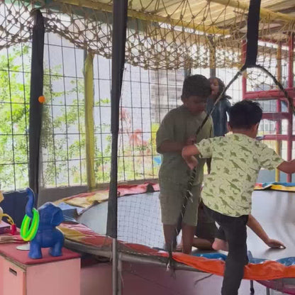 Children of various ages enjoy a vibrant indoor play area with a trampoline, ball pit, climbing structures, and a table for activities. The atmosphere is lively and playful, with kids actively engaged in jumping, climbing, and participating in a science experiment. Colorful soft blocks and play equipment create a stimulating environment for fun and learning.