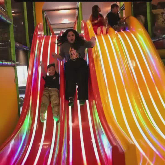 A child and an adult enjoy a vibrant indoor playground with colorful slides and a ball pit. Children of various ages are seen playing on a zip line, trampoline, and go-karts, creating a lively and fun atmosphere. The playground features exciting equipment like a tunnel slide and a climbing wall with a monkey theme.