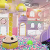 A vibrant indoor playground with a ball pit, slides, and a carousel offers a fun-filled experience for young children. The colorful and whimsical environment, filled with playful decorations and soft surfaces, creates a cheerful and engaging atmosphere. Children of various ages can be seen enjoying a range of activities, from sliding and playing in the ball pit to engaging in imaginative play at a toy kitchen and makeup station.