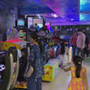 Children of various ages enjoy a vibrant arcade and play area, with attractions like racing games, a ball pit, and a climbing wall. The space is brightly lit with colorful lights and decorations, creating a lively and fun atmosphere. Families are present, with adults supervising and children actively engaged in play.
