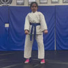 Young children in martial arts uniforms practice kicks and punches in a dojo with blue mats and a blue backdrop. An instructor holds pads for them to strike, while other children sit and watch. The atmosphere is focused and energetic.