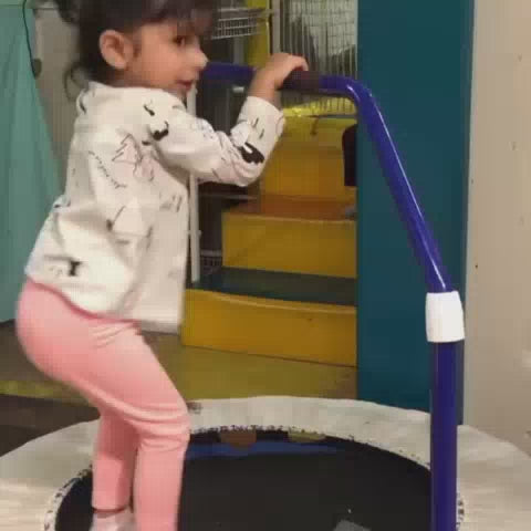 Young children joyfully play in a vibrant indoor playground filled with colorful slides, a ball pit, and soft climbing structures. Toddlers and preschoolers navigate the soft play area, some bouncing on a trampoline, while others slide down a bright yellow chute. The atmosphere is energetic and fun, with children of various ages actively engaged in play.