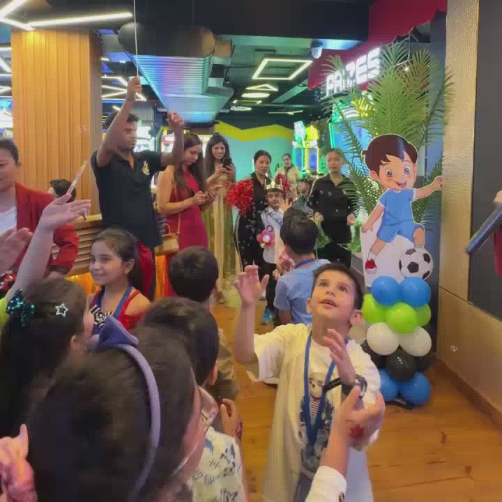 A lively birthday party unfolds at an arcade, with children of various ages excitedly playing and celebrating. Confetti fills the air as balloons float above, creating a festive atmosphere. The arcade's colorful lights and game machines provide a vibrant backdrop to the joyous occasion.