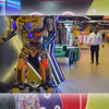 A vibrant arcade buzzes with activity as children and adults enjoy various games. Families engage in bowling, air hockey, and VR experiences, creating a lively and fun atmosphere. The setting is a brightly lit entertainment center filled with colorful arcade machines and attractions.