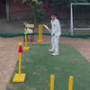 A young boy practices cricket on a grassy field with yellow wickets and cones. An adult supervises, offering guidance. The atmosphere is one of focused learning and skill development.