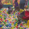 Children of various ages enjoy a vibrant indoor playground filled with colorful balls and slides. Some kids slide down a yellow chute, while others climb and swing. A young child plays with a toy cash register, simulating a shopping experience. The atmosphere is lively and playful, with children actively engaged in fun activities.
