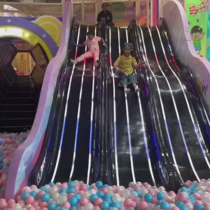Children of various ages enjoy a vibrant indoor playground filled with colorful ball pits, a thrilling slide, and interactive games. The atmosphere is lively and playful, with attractions like a trampoline, a cow-milking station, and swings adding to the fun.