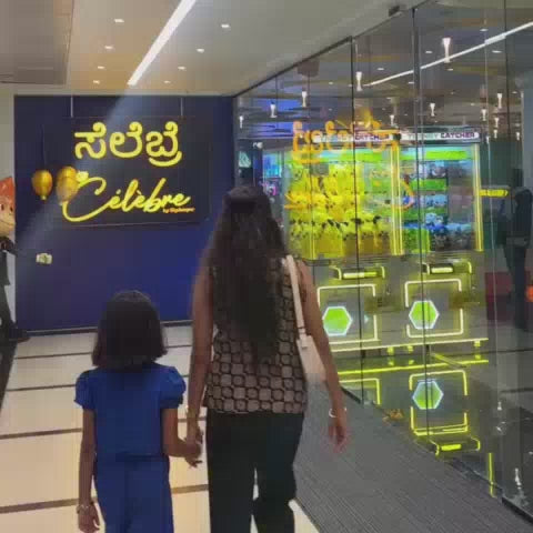 A family enjoys a vibrant arcade, with a young girl playing a motorcycle racing game and her mother trying her luck at a basketball arcade game. The brightly lit space, filled with colorful game machines and plush toys, creates a lively and fun atmosphere for all ages.