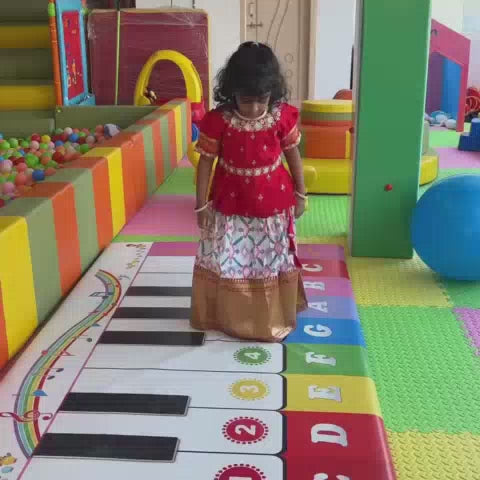Children of various ages enjoy a vibrant indoor playground filled with colorful ball pits, slides, a climbing wall, and a giant piano mat. The atmosphere is lively and playful, with kids actively engaging in climbing, sliding, and swinging.