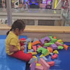 Children of various ages enjoy a vibrant indoor playground filled with soft blocks, ball pits, and slides. The atmosphere is lively and playful, with kids actively engaged in climbing, sliding, and interacting. The colorful equipment and netting create a safe and stimulating environment for active fun.