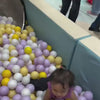A toddler joyfully plays in a ball pit filled with purple, yellow, and white balls. They then slide down a pink tube slide and a green slide. Later, the toddler interacts with a toy building set and enjoys a swing. Another young child is seen jumping on a trampoline. The atmosphere is lively and playful, suggesting a children's indoor play area.