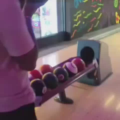 A group of young adults enjoys a lively arcade, with one person bowling and others playing a basketball arcade game. The atmosphere is energetic, with neon lights and colorful decorations enhancing the fun.
