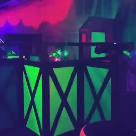 In a dimly lit laser tag arena, players of various ages navigate a neon-lit obstacle course filled with glowing green and pink structures. They wear illuminated vests and wield laser guns, engaging in a fast-paced game under blacklights. The atmosphere is energetic and exciting, with fog adding to the immersive experience.