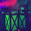 In a dimly lit laser tag arena, players of various ages navigate a neon-lit obstacle course filled with glowing green and pink structures. They wear illuminated vests and wield laser guns, engaging in a fast-paced game under blacklights. The atmosphere is energetic and exciting, with fog adding to the immersive experience.