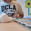A child, likely young, is engrossed in painting at a table, surrounded by art supplies like watercolors and brushes. The scene appears to be a home or classroom, with colorful decorations in the background. The atmosphere is one of focused creativity and quiet concentration.