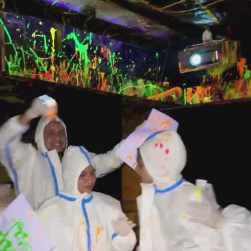 A group of young adults enjoys a vibrant, neon-lit paint party, splattering each other and the walls with glowing paint. They wear protective suits and gloves, creating a playful and energetic atmosphere. The room is a canvas of colorful paint splatters, with blacklights illuminating the scene.