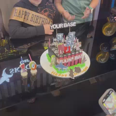 A birthday celebration at a gaming cafe features a cake with dinosaur decorations and "Happy Birthday" toppers. Children and teens are seen playing video games on multiple screens, some wearing headphones, creating a lively and fun atmosphere. The cafe is decorated with balloons and has a "Skycrest Gaming Cafe" sign.