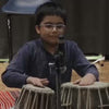A young boy plays the tabla in a performance setting. He is focused and engaged, with a microphone positioned to capture the sound. The atmosphere appears to be a formal recital or concert.
