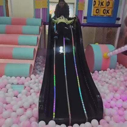 A young child enjoys a vibrant indoor playground, sliding down a black slide into a ball pit, playing at a pretend grocery store, and riding a toy car. Another child joins in the fun, interacting with a ball-dropping attraction and a carousel. The atmosphere is playful and engaging, with colorful equipment and soft flooring creating a safe and stimulating environment for young children.