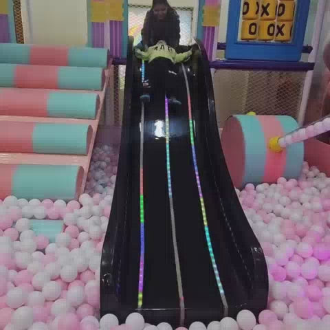 A young child enjoys a vibrant indoor playground, sliding down a black slide into a ball pit, playing at a pretend grocery store, and riding a toy car. Another child joins in the fun, interacting with a ball-dropping attraction and a carousel. The atmosphere is playful and engaging, with colorful equipment and soft flooring creating a safe and stimulating environment for young children.