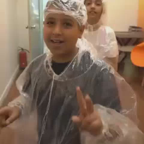 Children and adults are engaged in a colorful paint-splattering activity, possibly a "Paint it Wild" event. They are wearing protective gear like plastic ponchos and hairnets. The atmosphere appears joyful and energetic, with vibrant paint colors creating a lively environment.