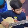 A young boy plays an acoustic guitar in a room filled with guitars and keyboards. Another boy plays a keyboard in the background, while a third boy is being instructed on guitar. The atmosphere appears to be a music lesson or practice session.