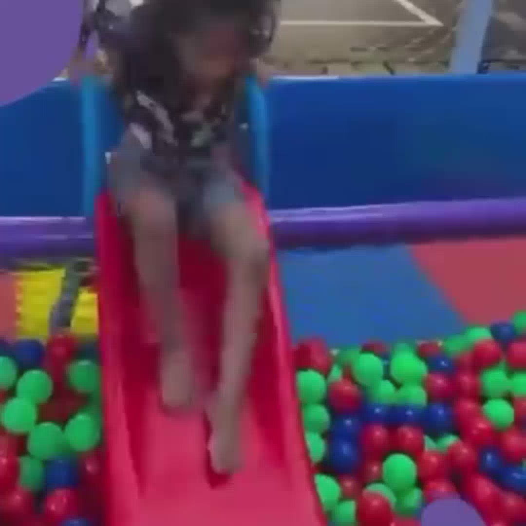Children of various ages enjoy a vibrant indoor playground filled with colorful ball pits, slides, and a climbing wall. The atmosphere is lively and playful as kids slide, climb, and tumble amidst a sea of red, green, and blue balls.