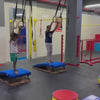 Two children, likely young, are engaged in a gymnastics-like activity in a brightly lit indoor gym. They are using various pieces of equipment, including rings, mats, and elevated platforms, to perform jumps and rolls. The atmosphere appears energetic and playful as they navigate the obstacle course.