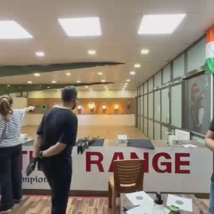 At a shooting range, individuals of various ages practice with rifles and pistols. The indoor environment features targets downrange and bright overhead lighting. The atmosphere appears focused and engaged as participants aim and shoot.