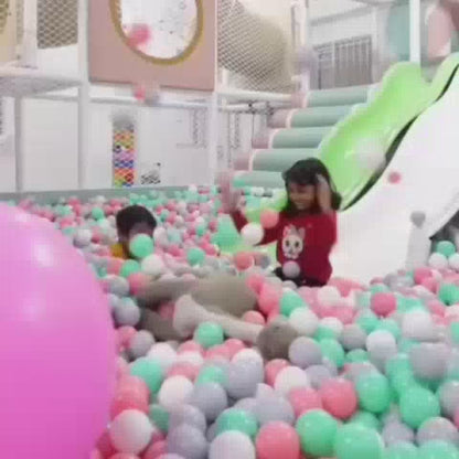 Children of various ages enjoy a vibrant indoor play area filled with colorful balls, slides, and climbing structures. The atmosphere is lively and playful, with kids engaging in activities like sliding, climbing, and building. The environment is designed for fun and exploration, featuring a ball pit, slides, and a rainbow-themed backdrop.
