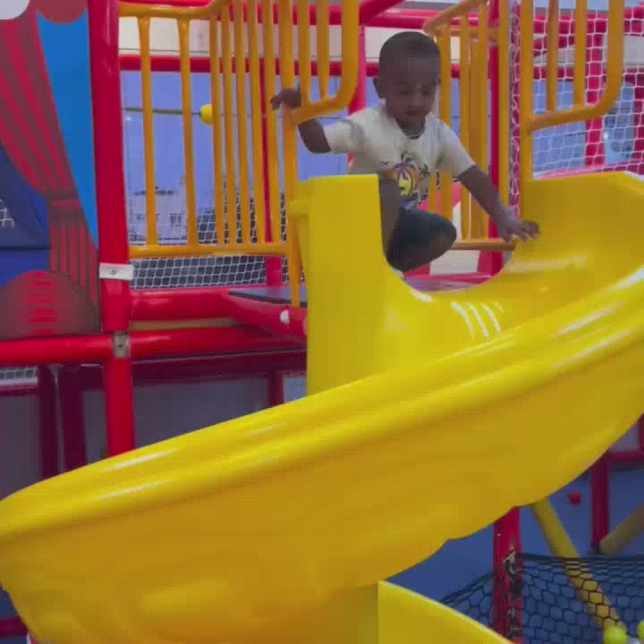 A young child gleefully navigates a vibrant indoor playground, sliding down a bright yellow slide and climbing soft, colorful steps. The environment is a child's paradise, filled with a ball pit and a trampoline, creating an atmosphere of pure fun and energetic play.