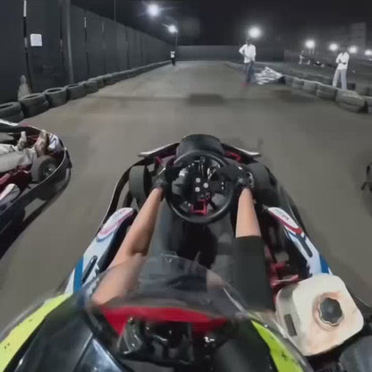 A group of young adults races go-karts at night on an outdoor track illuminated by bright lights. The atmosphere is energetic and competitive as drivers navigate the turns, with spectators watching from the sidelines. The go-karts are low-profile, open-wheeled vehicles, and the track is lined with tires for safety.
