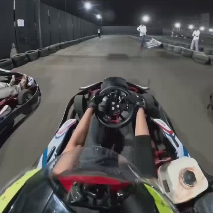A group of young adults races go-karts at night on an outdoor track illuminated by bright lights. The atmosphere is energetic and competitive as drivers navigate the turns, with spectators watching from the sidelines. The go-karts are low-profile, open-wheeled vehicles, and the track is lined with tires for safety.