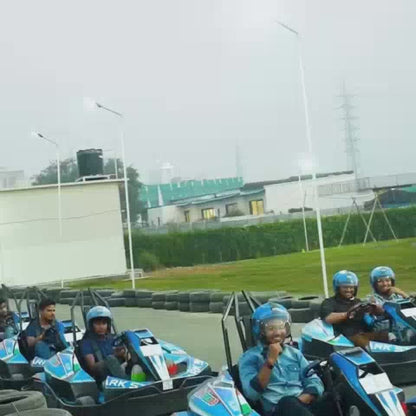 A group of people, including children and adults, enjoy various attractions at an indoor amusement park. The park features go-karts, a ball pit, a climbing structure, trampolines, and a basketball hoop area. The atmosphere appears lively and fun, with bright colors and energetic activities.