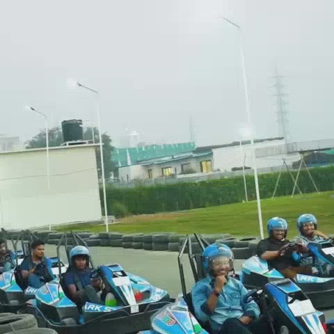 A group of people, including children and adults, enjoy various attractions at an indoor amusement park. The park features go-karts, a ball pit, a climbing structure, trampolines, and a basketball hoop area. The atmosphere appears lively and fun, with bright colors and energetic activities.