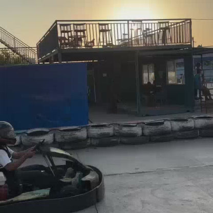Go-karting enthusiasts of various ages race around a track lined with tires, under a sunset sky. The atmosphere is energetic, with colorful graffiti art adorning the surrounding buildings and a prominent "Flying Go-Karts" banner.