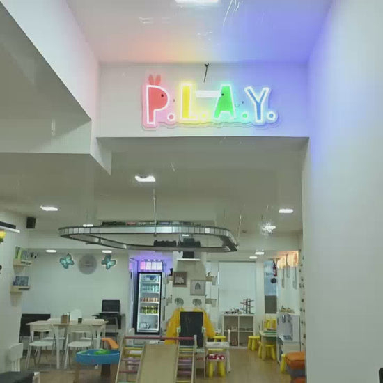 This vibrant play space, likely for young children, features a colorful "P.L.A.Y." sign, a miniature train track, a slide, and a world map mural. The environment is bright and engaging, with shelves stocked with books and toys, creating a fun and stimulating atmosphere.